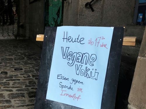 Ein Holzaufsteller vor einem Hof, auf dem ein handgemaltes Papierplakat hängt, auf dem steht: "Heute ab 17.30 Uhr Vegane VoKü. Essen gegen Spende"